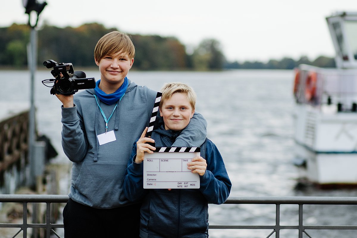 Zwei Jungen mit Kamera und Filmklappe stehen Arm um Arm und Lächeln - Impression des Filmworkshops der Kinderfilmuni in Neuruppin