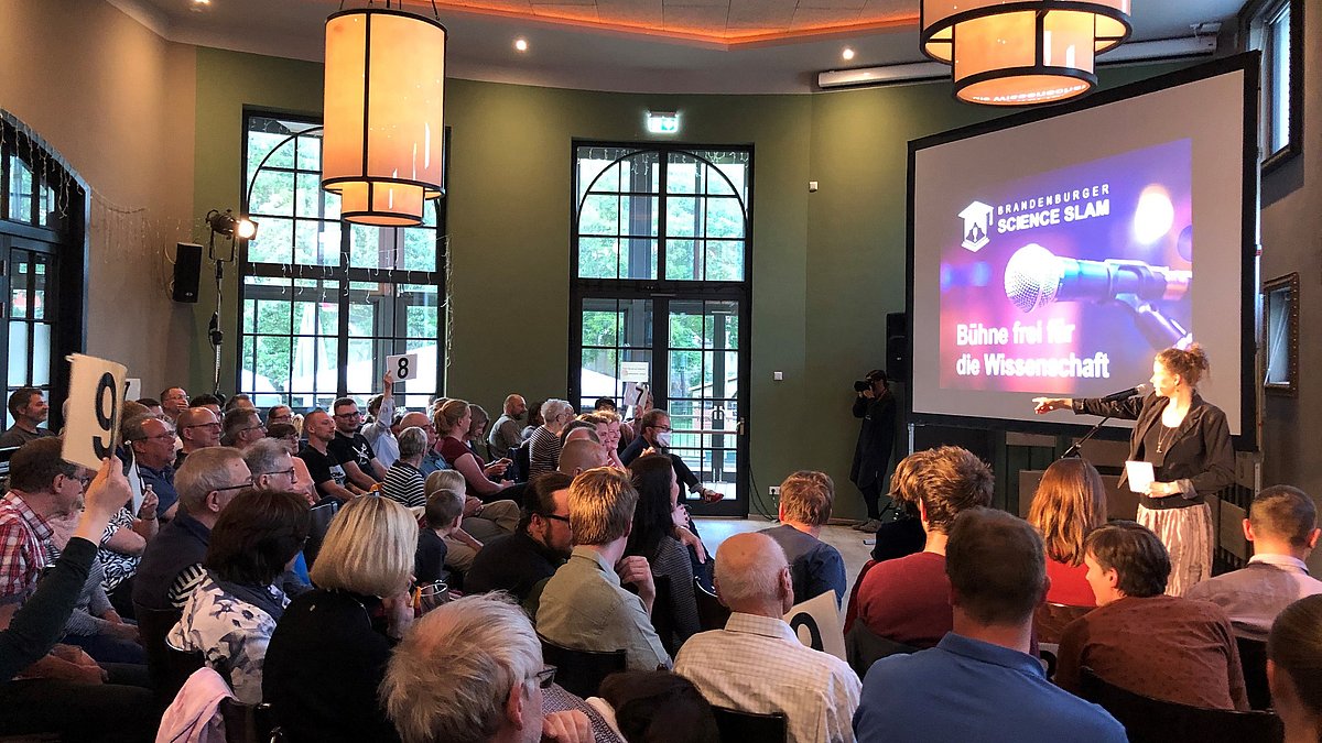 Veranstaltungsabend Science Slam 2022 im BrauWerk Schwedt/Oder. Das Publikum gibt Punktewertung ab eine Dame auf der Bühne zählt die Stimmen aus.