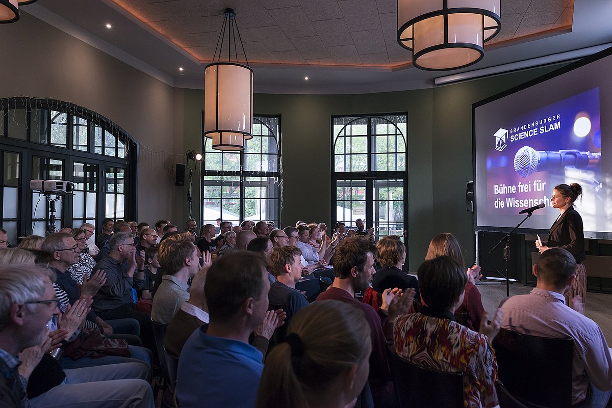 Ein voll besetzter Publikumsraum mit ca. 80 Personen. Vorne rechts sieht man eine Leinwand mit Titelbild des 1. Brandenburger Science Slams. Die Moderatorin spricht daneben in ein Mikrofon.