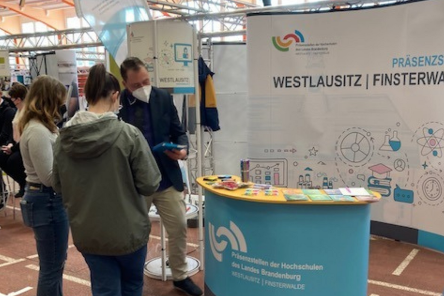 Messestand der Präsenzstelle auf einer Messe mit Beratungssituation zwischen Ansprechperson und zwei Gästen