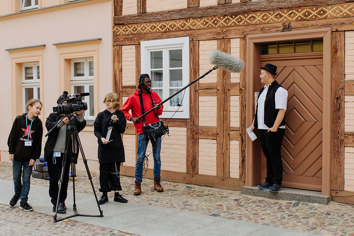 Situation aus einem Workshop der Kinderfilmuni in Neuruppin. Kinder an der Filmkamera, mit Mikrofon und Filmklappe filmen einen Jungen, der im Türrahmen steht