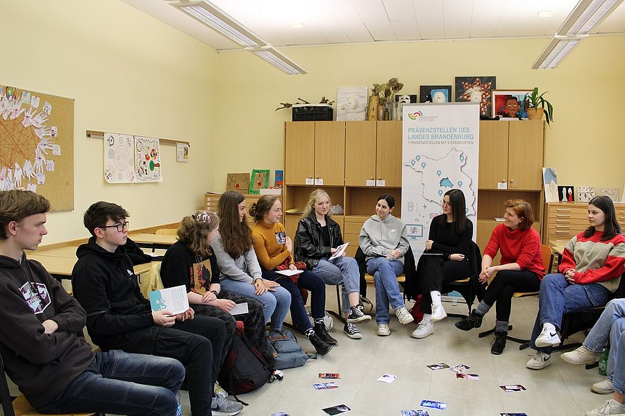 Schülerinnen und Schüler sitzen mit Workshopleiterin im Stuhlkreis und betrachten Fotos aus verschiedenen Berufsbereichen.