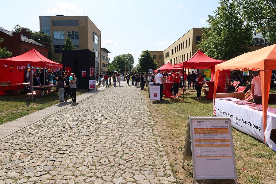 Bild auf dem Hochschulcampus mit den verschiedenen Ständen der Aussteller zum Tag der offenen Tür. Besucherinnen und Besucher gehen über den Campus und informieren sich an den Ständen. 