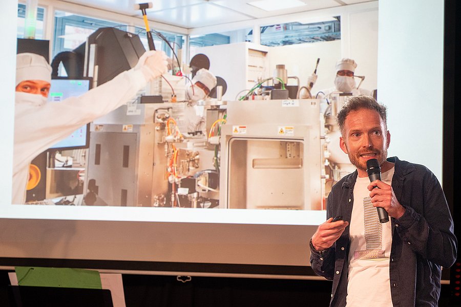 Sieger, Sebastian Schulze, vom Leibnit-Institut für innovative Mikroelektronik, beim letzten Brandenburger Science Slam in Finsterwalde. Im Hintergrund sieht man seine Präsentation mit einem Foto von ihm und seinen Kollegen im Labor in voller weißer Schutzkleidung.
