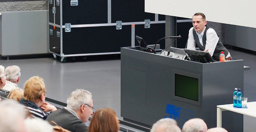 Blick in einen Hörsaal mit Sitzen in Treppenanordnung. Die Zuhörenden sind alle in einem fortgeschrittenen Alter und haben zum Teil graue Haare. Der Blick ist über ihren Rücken zum Dozenten gerichtet, der auf das Rednerpult gestützt ist.