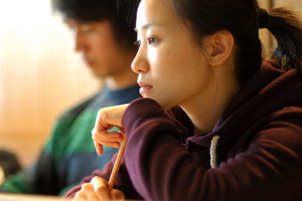 Eine sitzende Studierende lehnt ihre Hand und den Kopf auf einen Stift, den sie mit der zweiten Hand festhält. Neben ihr sitzt ein Kommilitone, der nur verschwommen dargestellt ist.