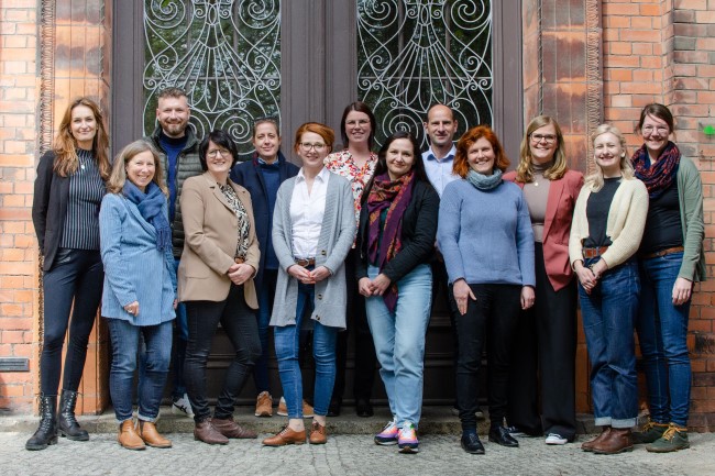Teamfoto von 13 Mitarbeitenden der Präsenzstellen der Hochschulen des Landes Brandenburg. Vor einem roten Backsteingebäude mit großer Eingangstür aus Holz mit Fenstern und verschnörkeltem Metallgitter davor.